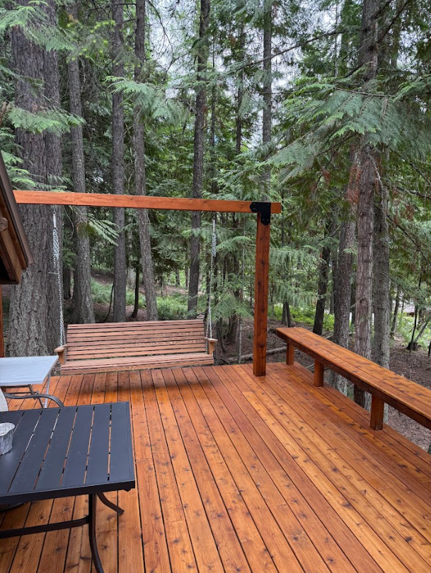 Wooden deck with swing, bench, and table surrounded by tall trees. The wood is stained a reddish-brown.