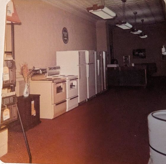 An old appliance store interior.  White fridges and stoves line the wall. Dark red flooring.