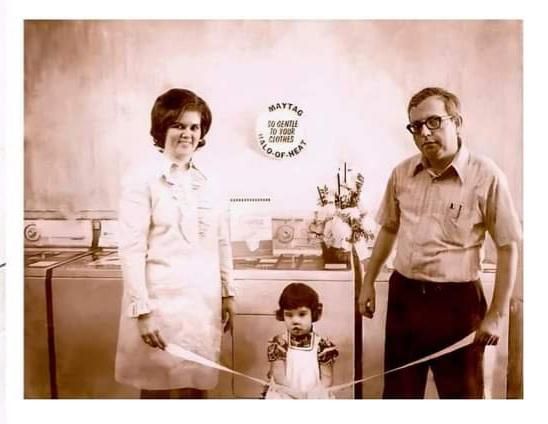 A family stands in a laundromat, holding a ribbon; mother in white, father in glasses, child seated.
