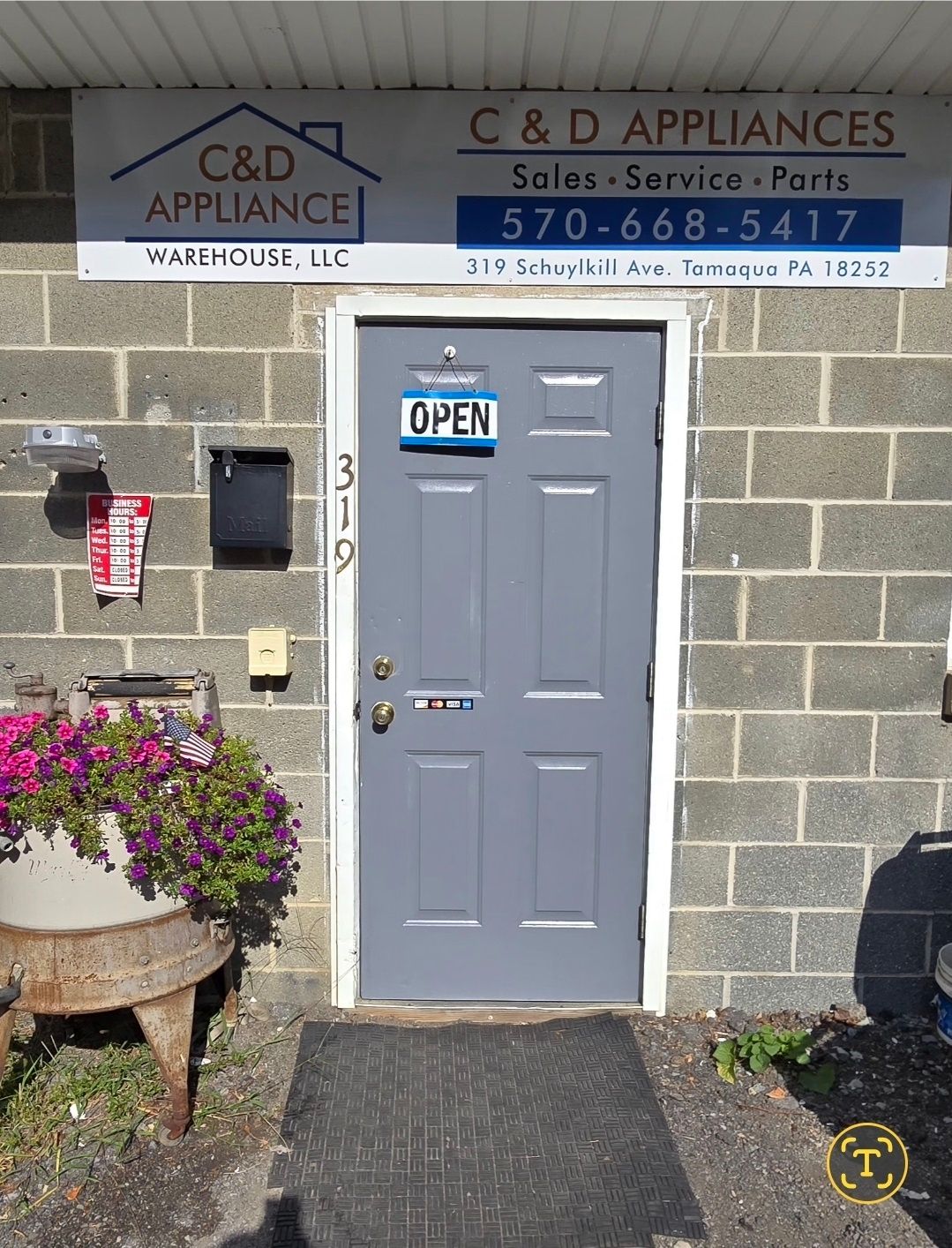 Gray door of C&D Appliances shop with open sign. Brick building, sign, mailbox, flowers.