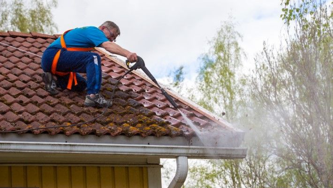Worker pressure-washing a red-tiled roof with a hose on a house