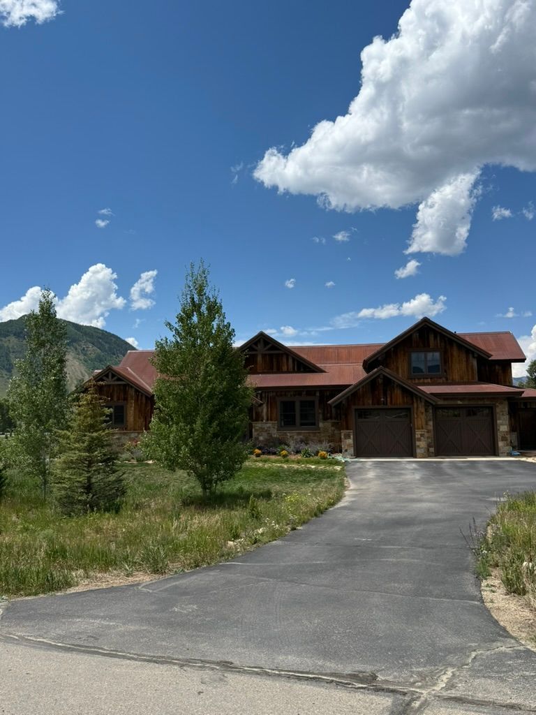 Large wood and stone house with driveway, set against a mountain backdrop and a blue sky with puffy white clouds.