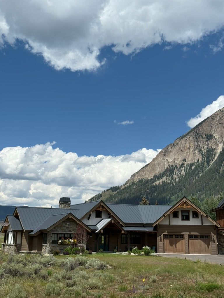 House with dark gray roof, beige exterior, set against a mountain and blue sky with clouds.