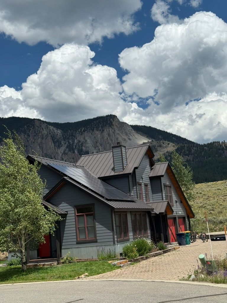 Gray house with solar panels, red doors, and mountains under a cloudy blue sky.