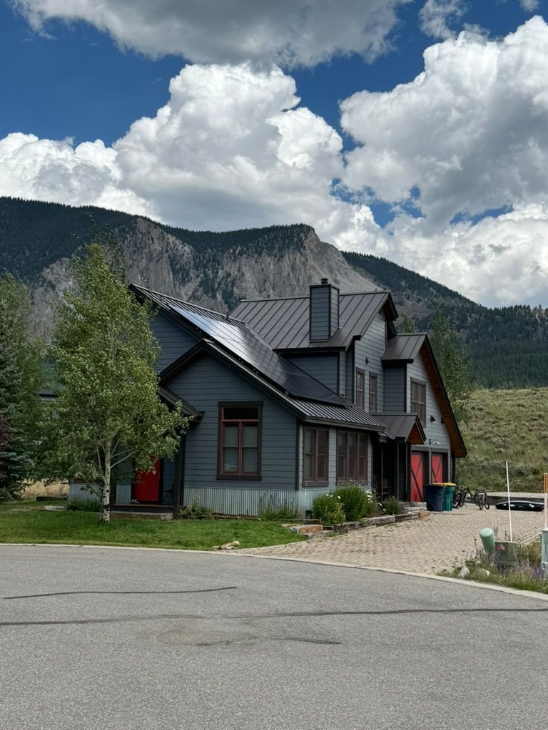 Gray house with dark roof, red door, against mountain, cloudy sky.