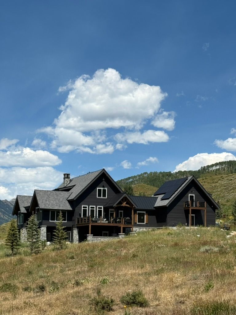 Dark-colored house with multiple gabled roofs, on a hillside under a blue sky with clouds.