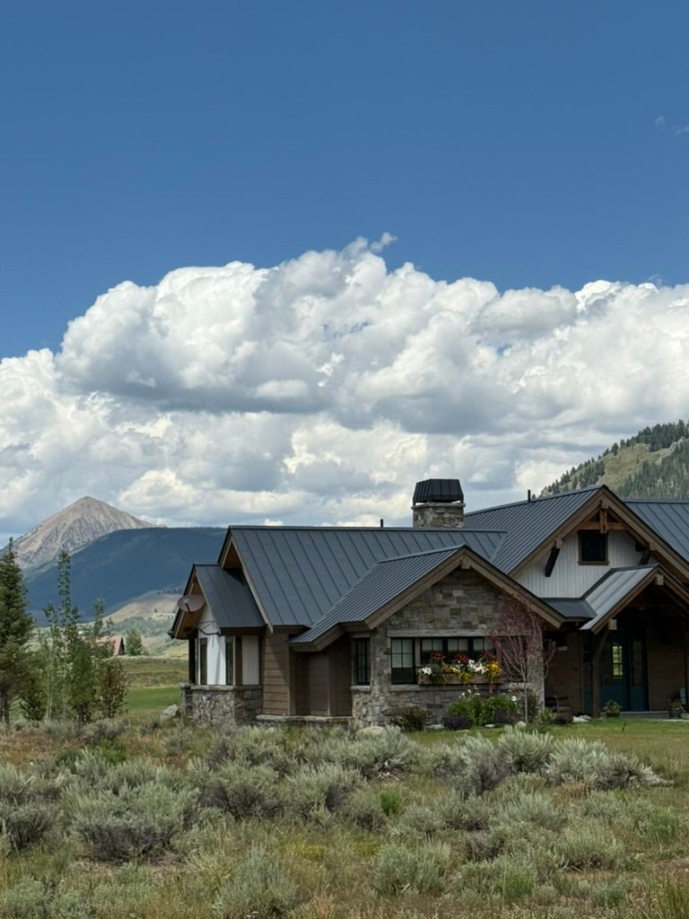 Stone and wood home with dark roof, nestled in front of mountains under a cloudy sky.