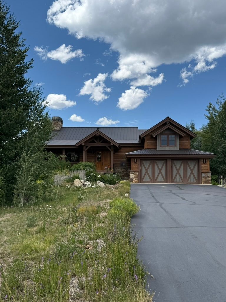 Log cabin home with dark roof, wooden garage doors, and a paved driveway under a blue sky with clouds.