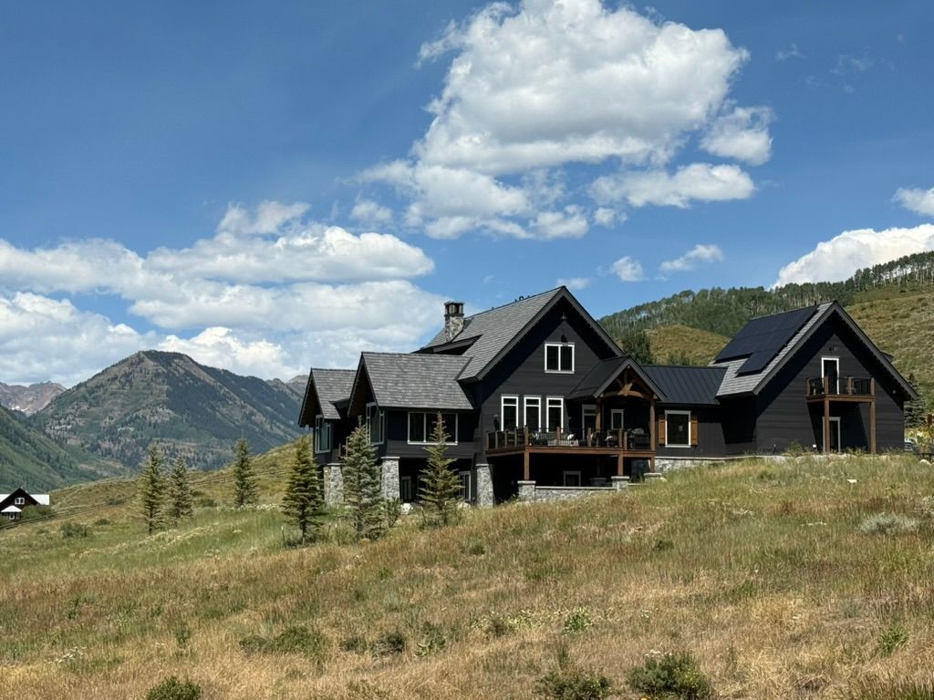 Dark house with multiple gables on a hillside with mountains and blue sky.