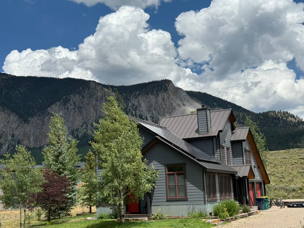 Gray house with dark roof against mountain and cloudy sky.