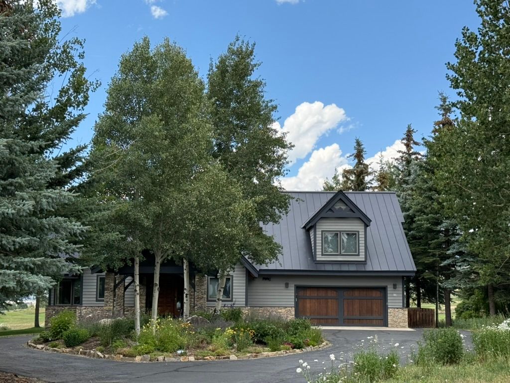 House exterior with driveway, trees, and blue sky. Gray siding and dark roof, wooden garage doors.