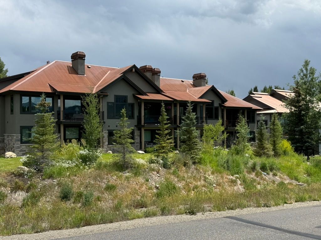Multi-unit, brown-sided building with rusty metal roof and stone accents, in a grassy, mountain setting.