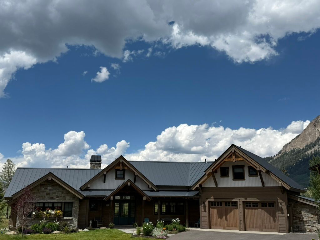 House with stone and wood facade under a blue sky with clouds, mountains in the background.