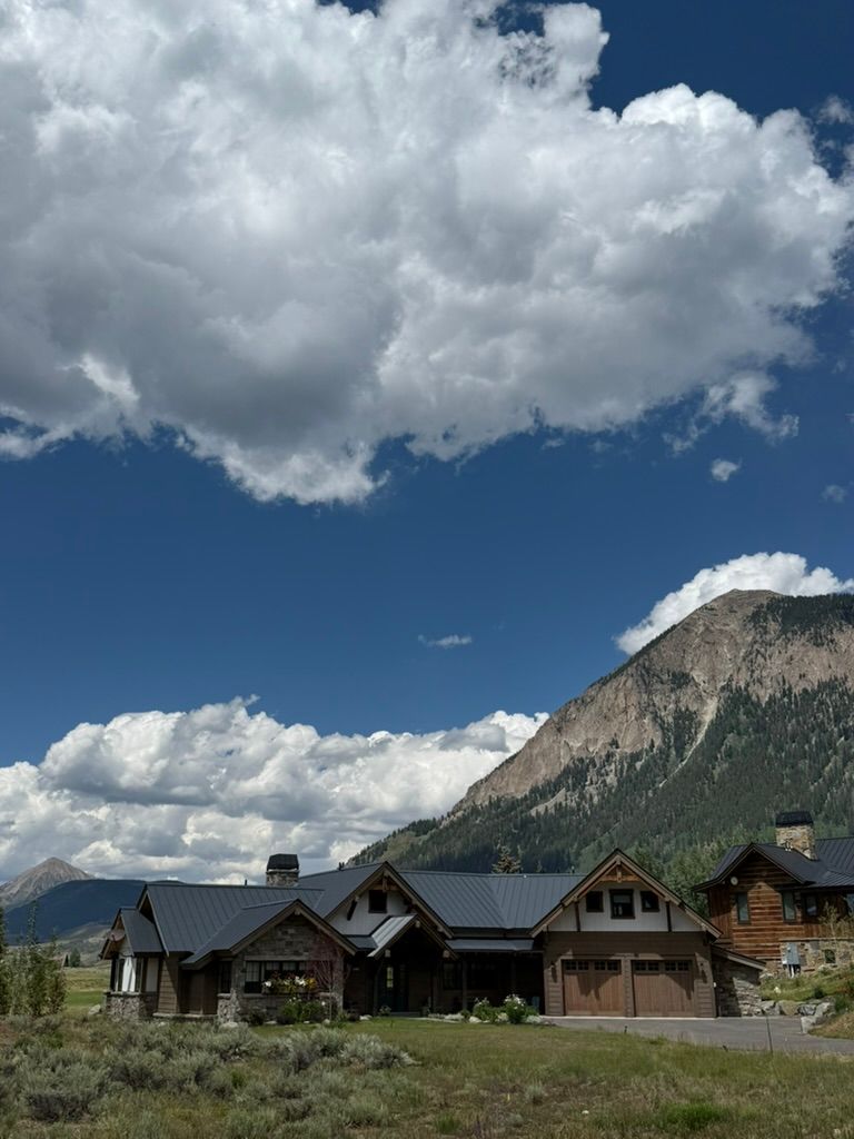 Mountain home with dark roof under blue sky, white clouds, and mountain background.