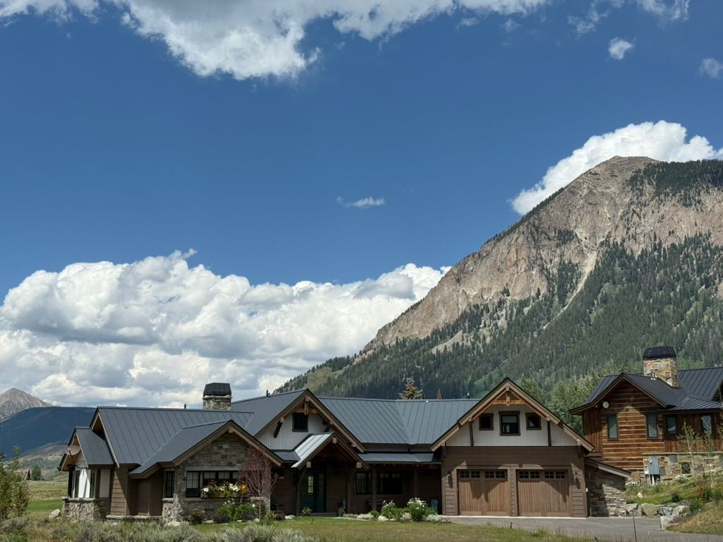 Houses with dark roofs and stone accents sit in front of a mountain under a blue sky with clouds.
