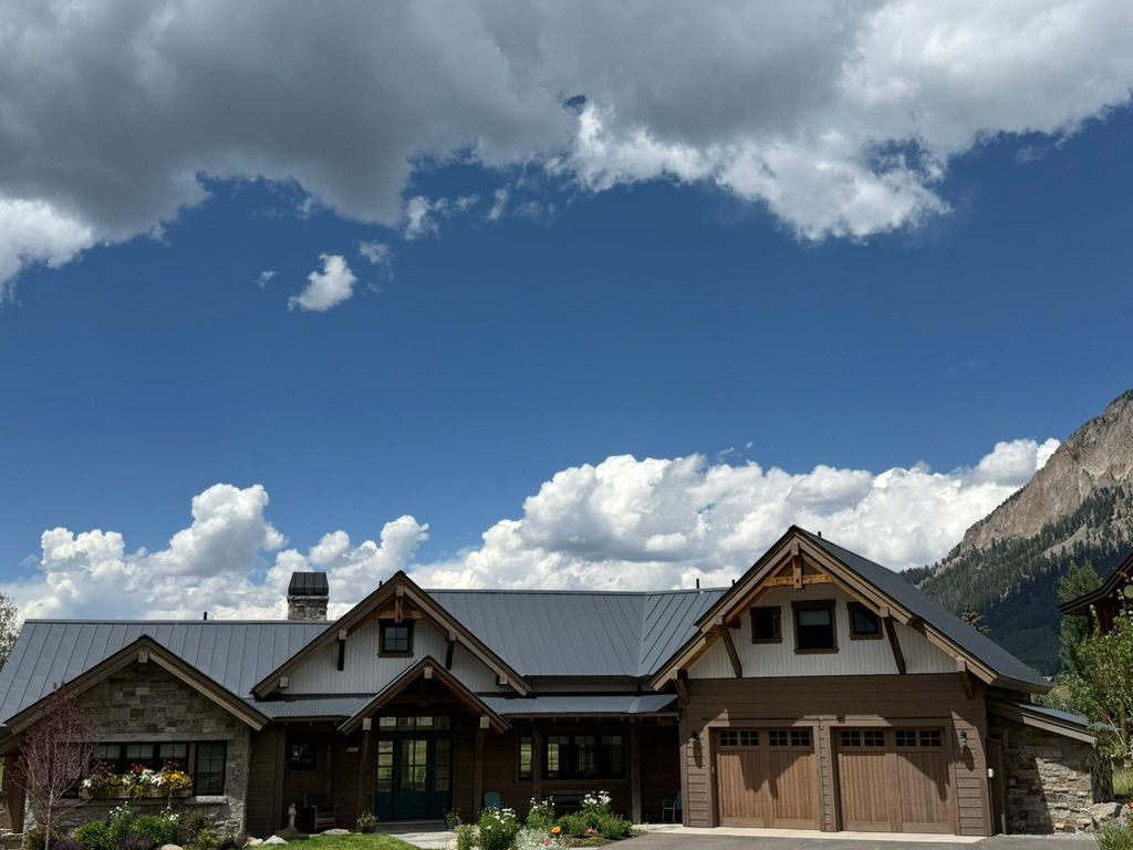 Large house with stone and wood facade under a blue sky with white clouds and mountain in the background.