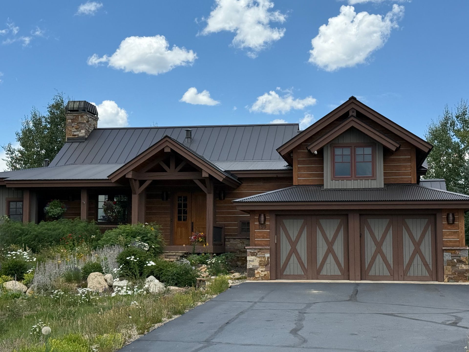 Log cabin-style house with brown wood, metal roof, stone accents, and two-car garage under a blue sky.