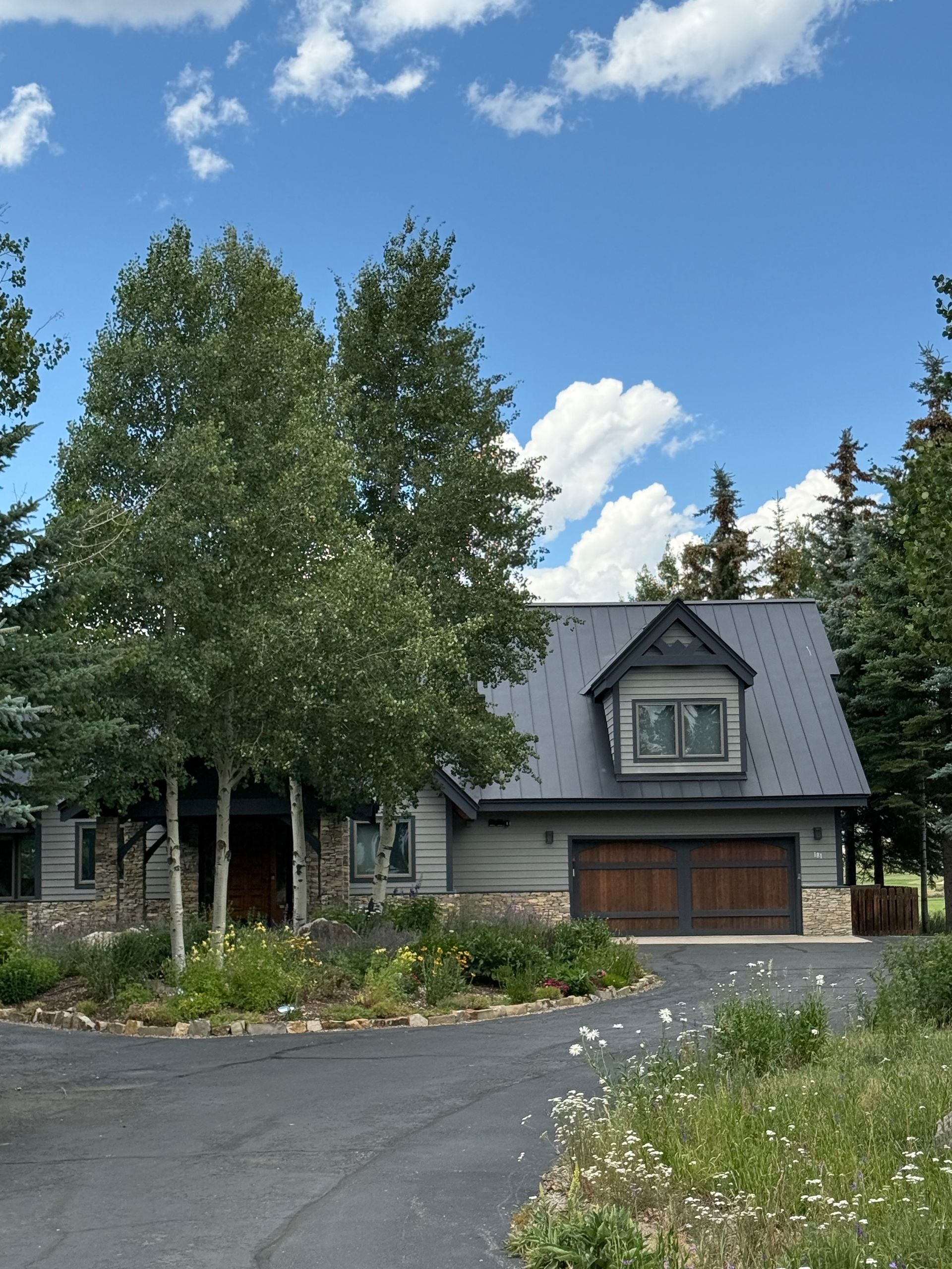 A gray house with a black roof and wooden garage doors. Two tall trees stand in front.