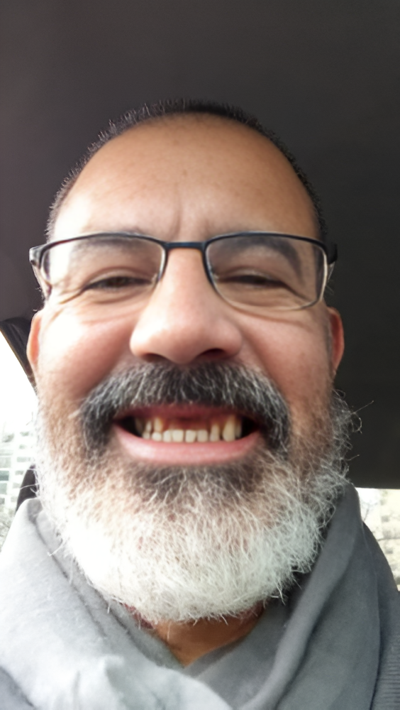 Hombre sonriente con gafas y barba gris, primer plano en interiores.