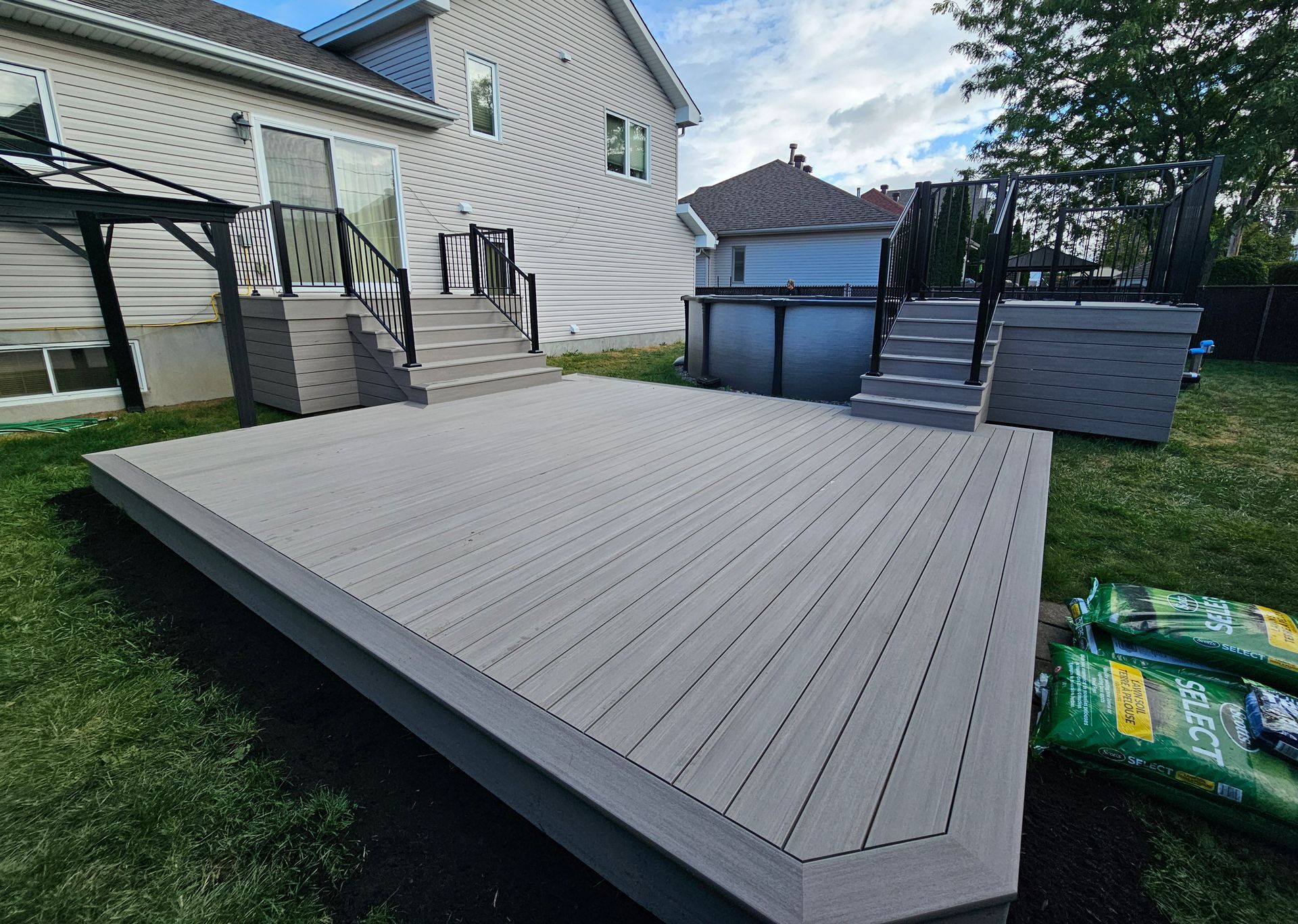Grande terrasse grise jouxtant une maison et une piscine hors sol dans un jardin, avec des sacs de jardinage sur la droite.