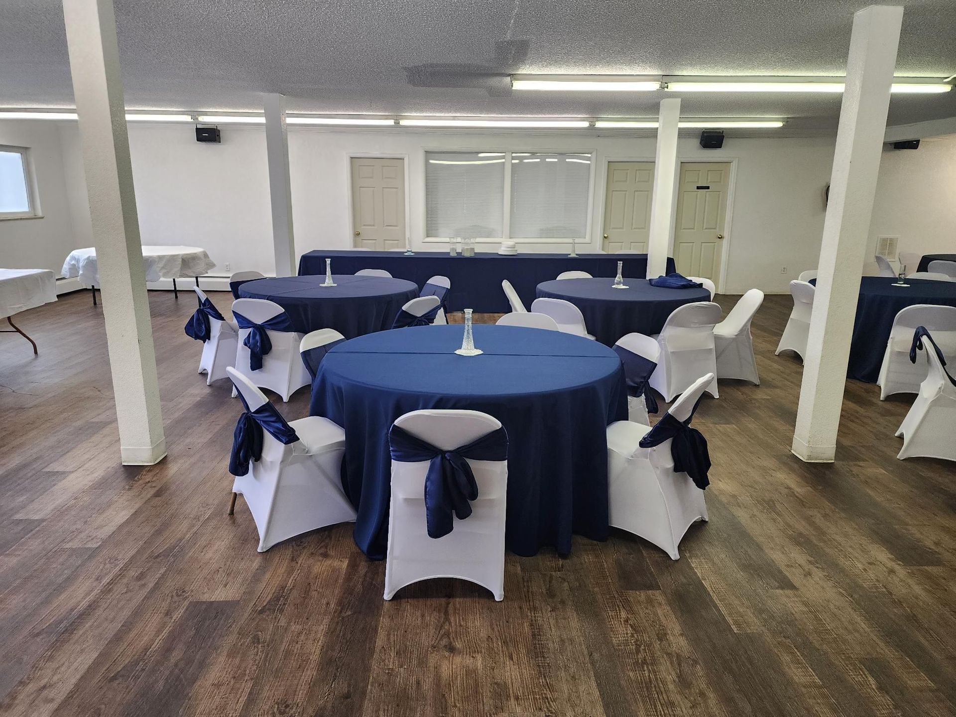 Room with round tables covered in navy blue linens, white chairs with navy blue sashes, and wooden floor.