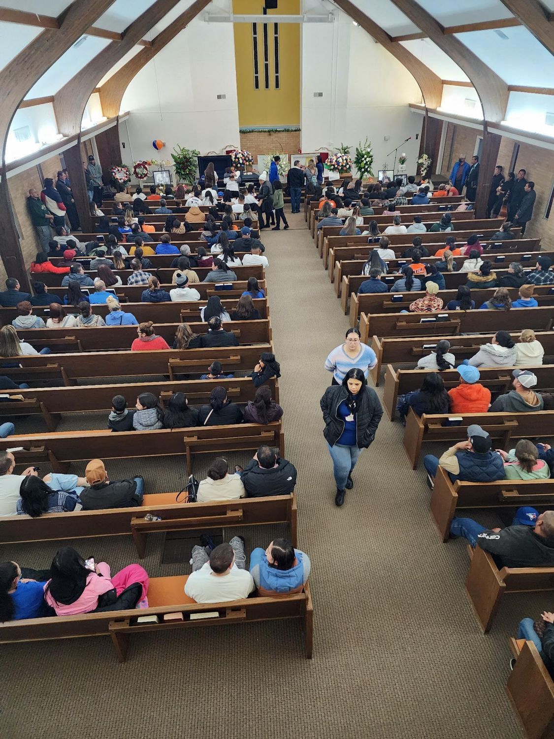 Church interior with many people seated in pews, facing the altar.  A person walks down the aisle.