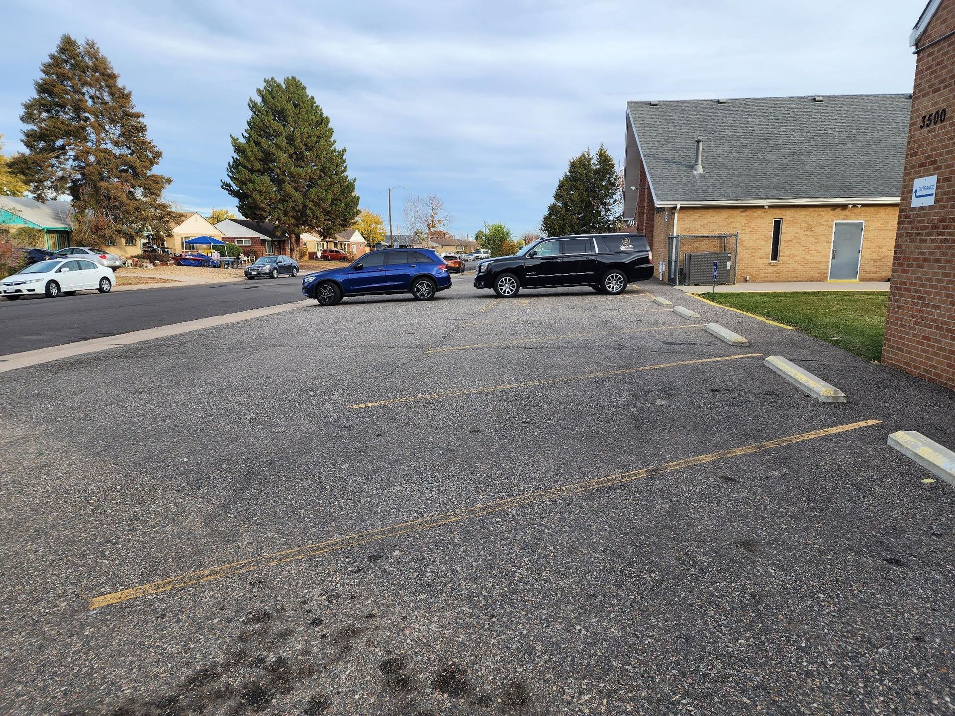 Cars parked on gravel in front of a building; a street and trees are in the background.