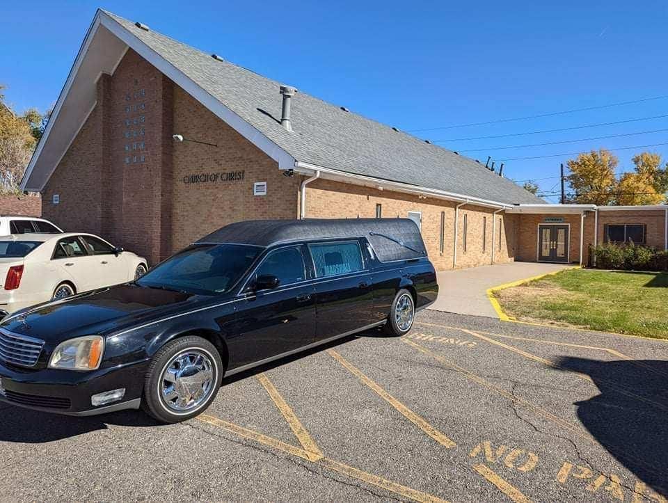 Black hearse parked outside a brick building with a sloped roof under a blue sky.