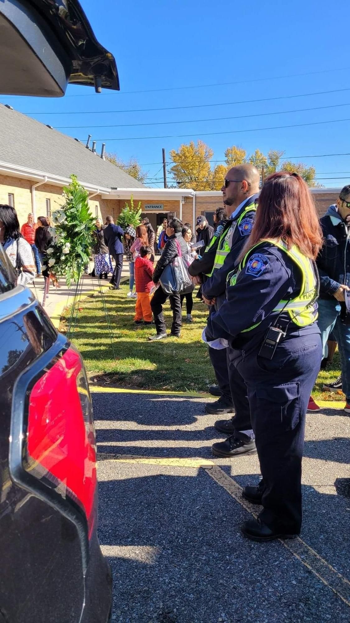 Police officers standing near a vehicle, facing a crowd outside a building on a sunny day.