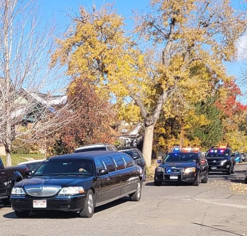 Black limousine escorted by police vehicles on a tree-lined street with autumn foliage.
