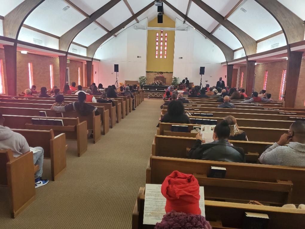 Inside a church, people sit in pews facing the altar. The space is well-lit with high ceilings.