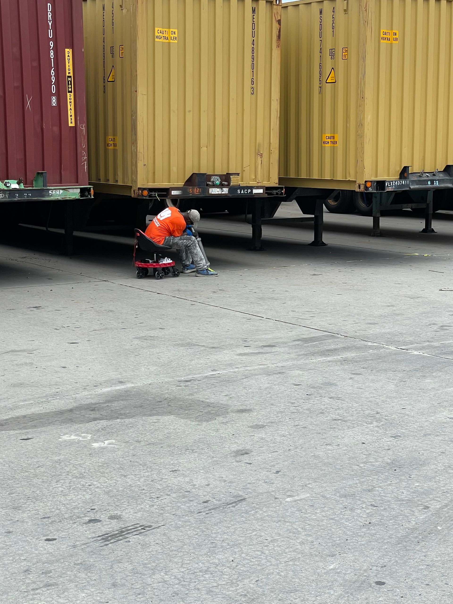 A man sits on the ground in front of a row of shipping containers