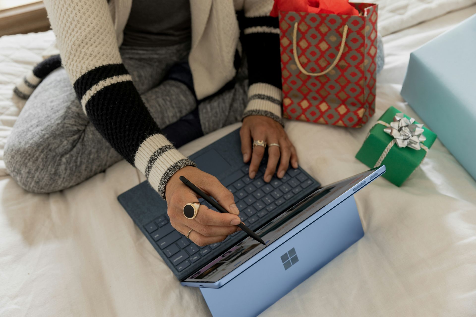 A person uses a stylus on a blue Microsoft Surface tablet, sitting on a bed with holiday gift bags 