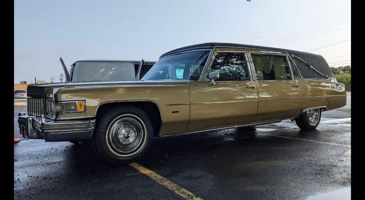 Gold vintage Cadillac hearse parked outdoors on wet pavement.