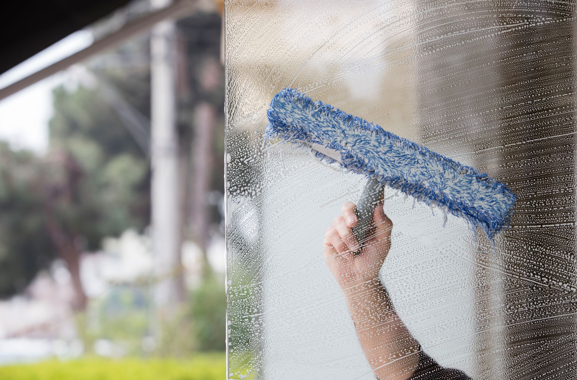 A hand uses a blue scrub brush to clean a glass surface covered in soap suds.