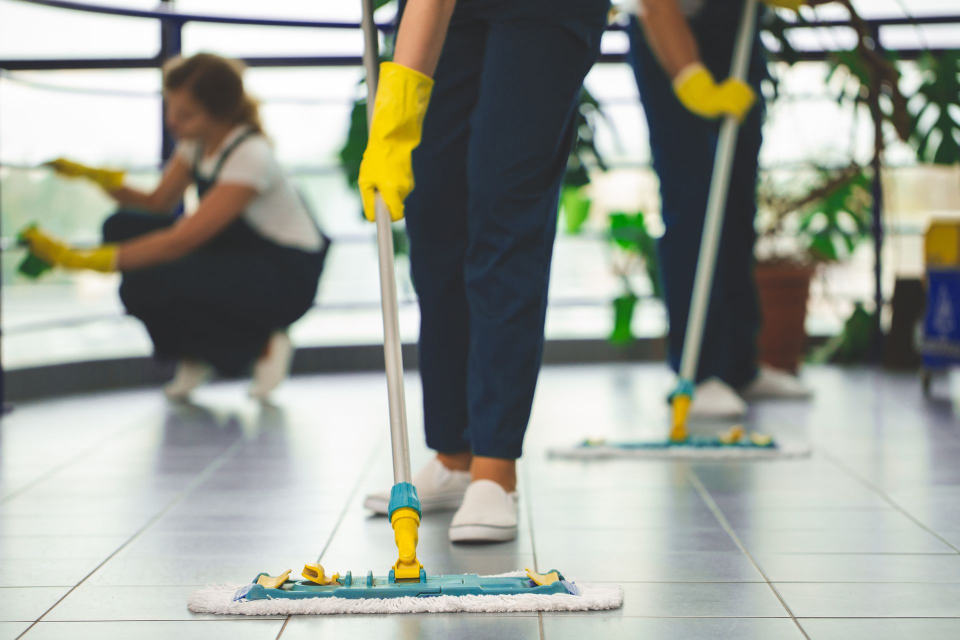 Three cleaners wearing yellow gloves work in an office setting, mopping floors and wiping a glass railing.