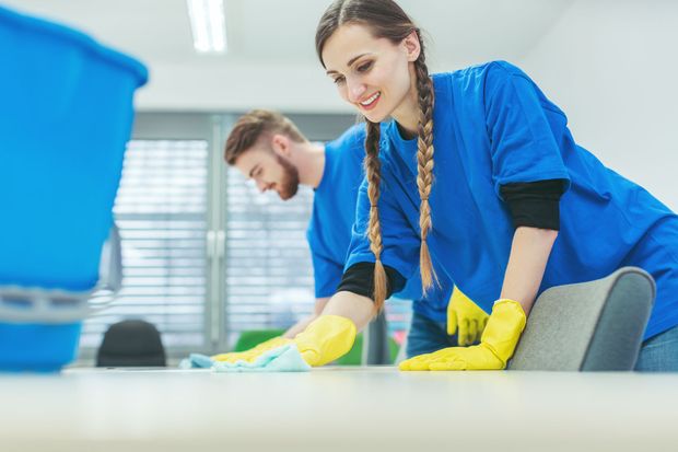 A cleaner wearing orange gloves and a uniform holds a bucket filled with various cleaning supplies in an office.