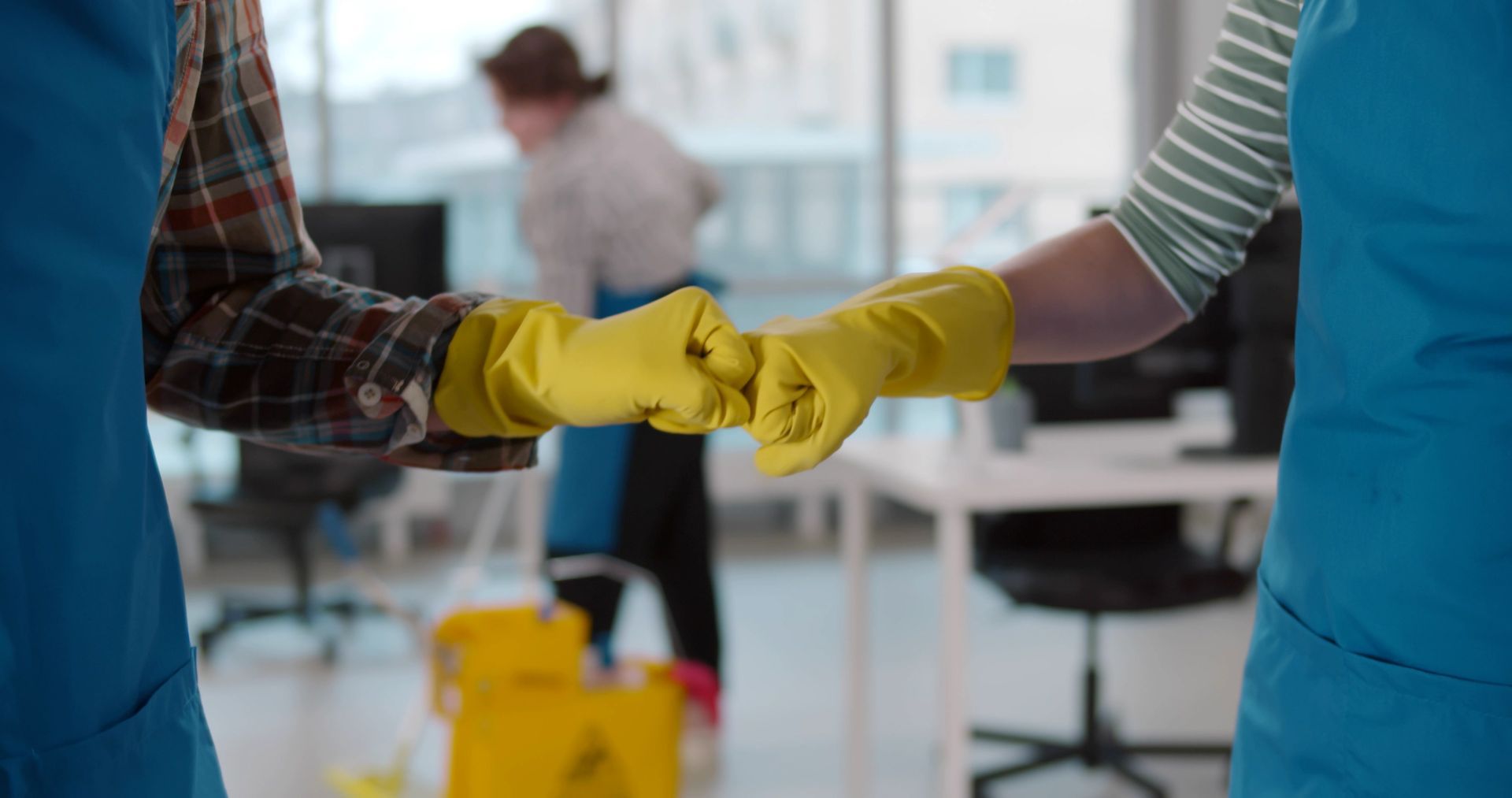 Two people in blue aprons and yellow rubber gloves perform a fist bump in an office.