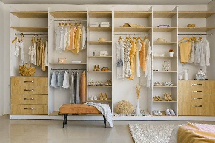 Built-in closet with beige shelves, hanging clothes, drawers, and bench. Soft neutral colors.