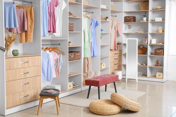 Walk-in closet with organized clothing, shelves, drawers, bench, and mirror. White and beige with pastel colors.