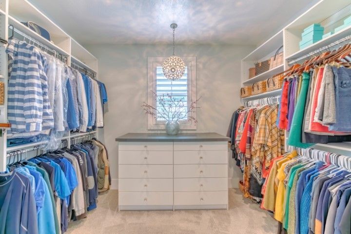 Walk-in closet with white shelves and clothing racks filled with clothes. A central drawer unit sits below a window.