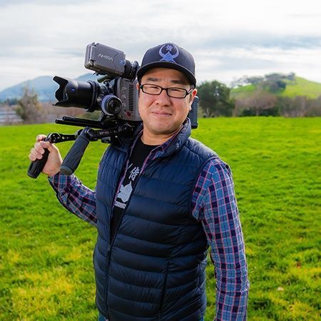 A man in glasses, holding a large camera on his shoulder outdoors, wearing a navy vest, plaid shirt, and baseball cap.