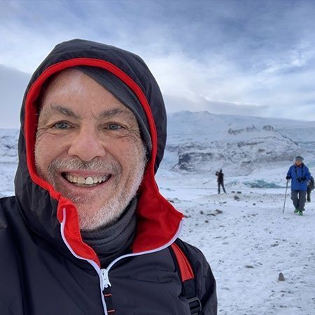 Man smiles in snowy landscape wearing black and red hooded jacket. Two figures hike in distance, near a glacier.