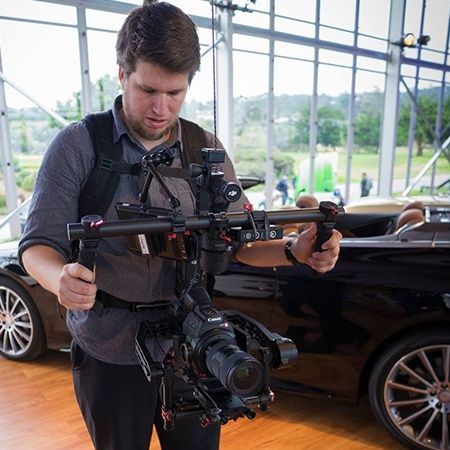 Man filming with a camera rig, appearing to focus. He stands indoors near a black car.