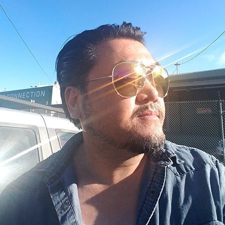 Man with sunglasses and a goatee, looking right, outdoors on a sunny day. He wears a denim shirt.