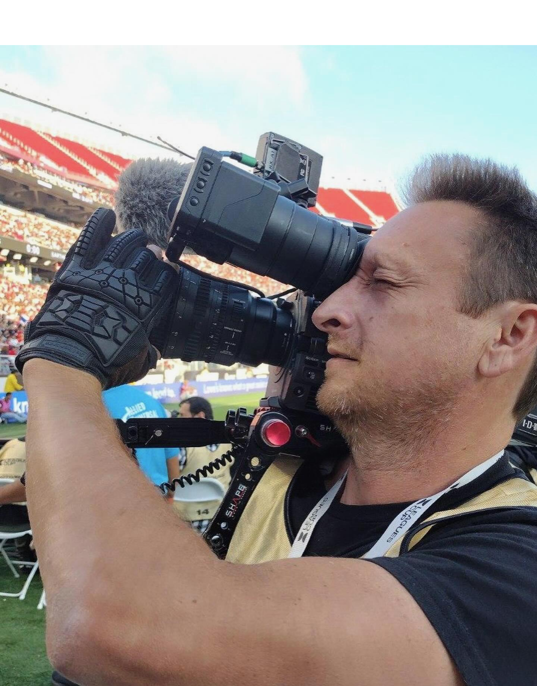 Cameraman wearing black gloves and a vest, filming at an outdoor stadium. He holds the camera to his eye, focused on the event.
