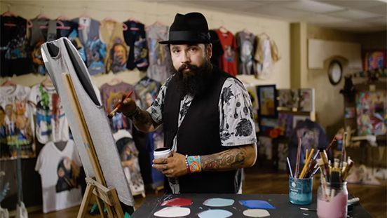 A bearded artist in a hat paints a surfboard in his studio, with colorful shirts hanging in the background.