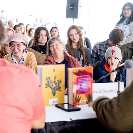 People listen at a book event. Two books on a table, faces smiling, microphones present.