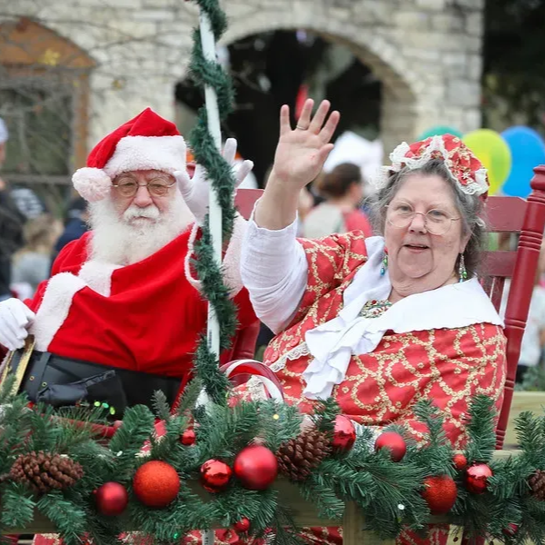 Santa and Mrs. Claus waving from a decorated sleigh. Red and white outfits, festive decor, outdoor setting.
