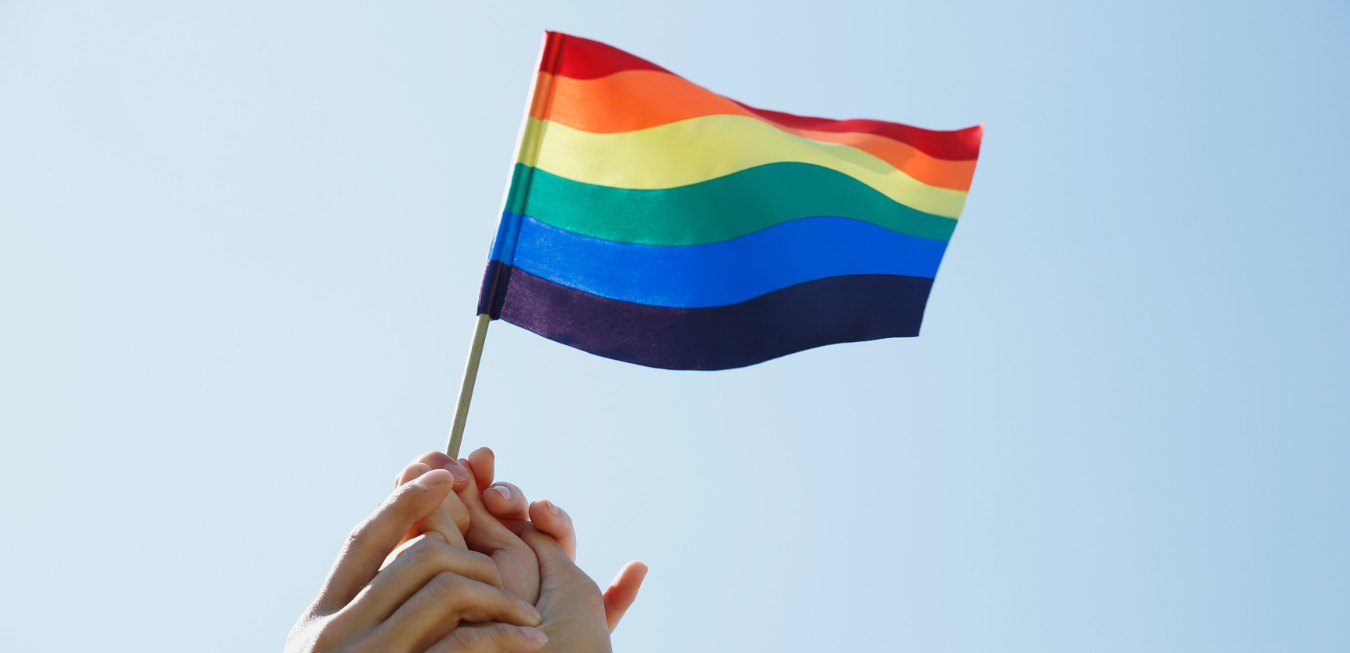 Hands holding a small rainbow pride flag against a clear blue sky.